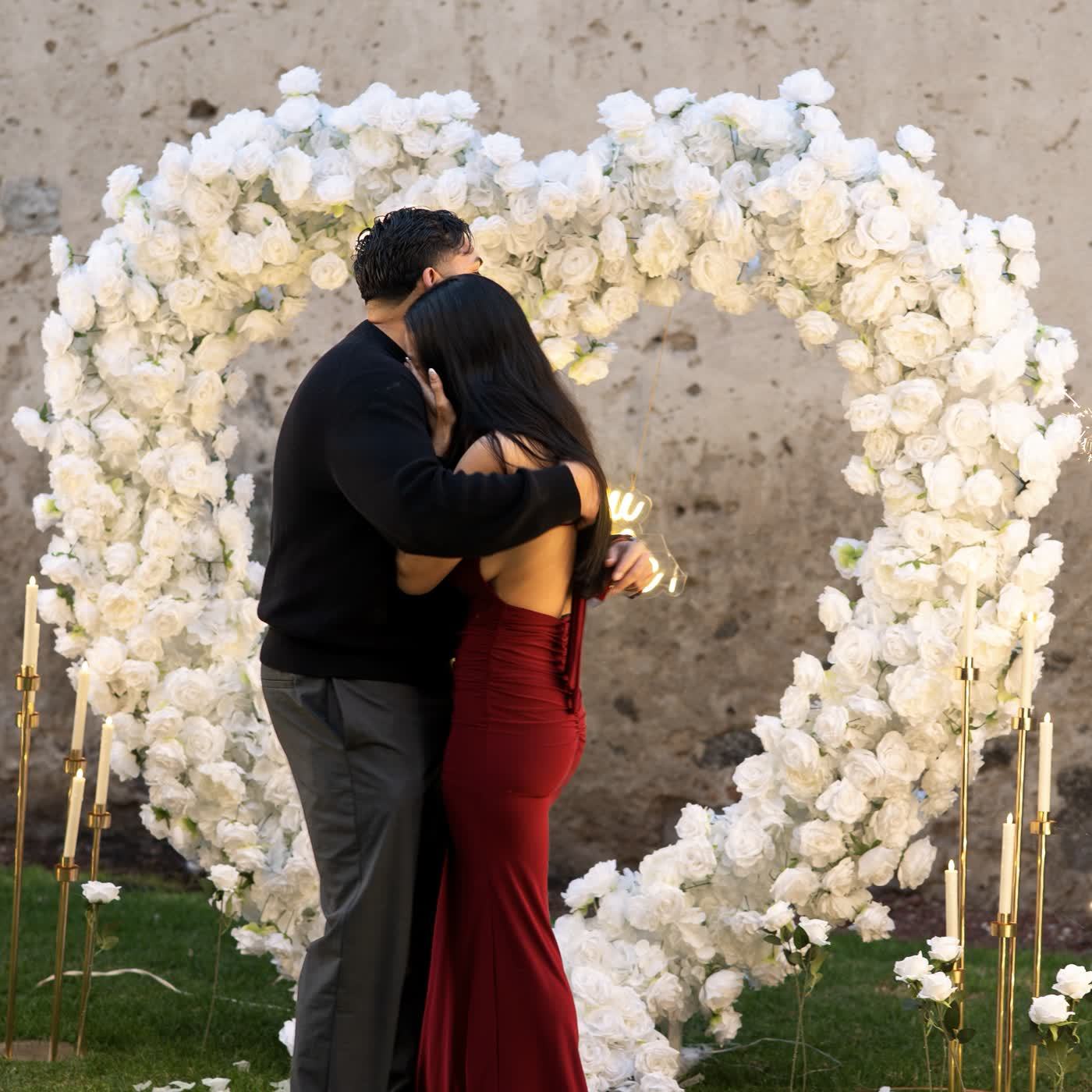 Pareja abrazada frente a corazón de rosas blancas en propuesta de matrimonio
