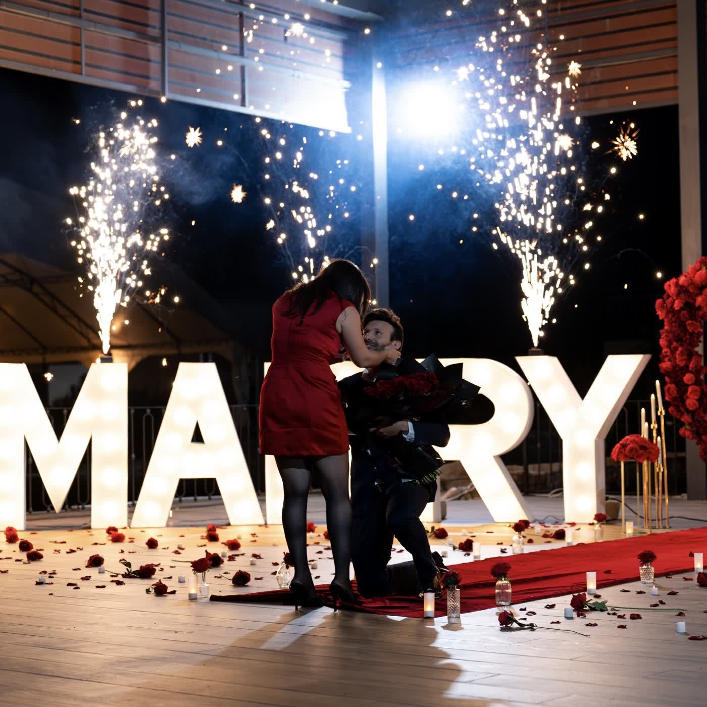 Letras gigantes iluminadas MARRY con fuegos artificiales y pareja abrazada